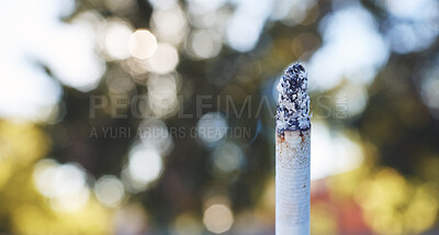 Closeup of a burning cigarette with an ashy tip against a blurred background outside. Tobacco is an addictive and harmful substance that can lead to health issues. Nicotine addiction can be a gateway