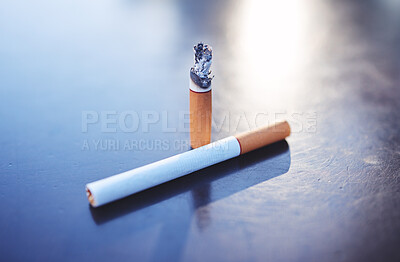 Above shot of two cigarettes, one lit and burning, on a table. Tobacco and nicotine are harmful and addictive substances that can lead to health issues in the future. Smoking is known to cause cancer