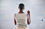 Black female looking at analytics, seo research, and statistics on a whiteboard strategizing the next move for her startup business. A motivated modern woman with a clear vision thinking and innovating