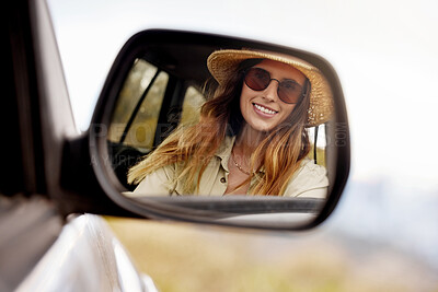 Reflection in the rear view mirror of a young beautiful woman\'s car while travelling to a vacation. Caucasian woman wearing sunglasses and a hat inside a car going on a roadtrip looking at her reflection and smiling while driving on the road