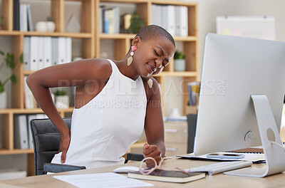 Young african american businesswoman suffering from back pain while sitting in a chair and using a computer in an office at work. Unhappy black woman looking uncomfortable while suffering back pain