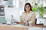 Portrait on one beautiful young mixed race businesswoman working on her computer in the office at work. Confident and successful female entrepreneur of indian ethnicity working on a desktop in her corporate workplace looking motivated and happy.