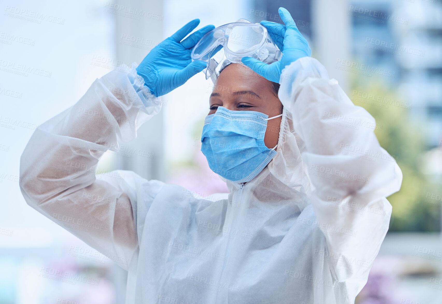 Buy stock photo Young doctor removing her protective goggles during the covid pandemic at the hospital. Medical professional wearing gloves, safety suit and a mask in her office at work. Healthcare worker in a clinic