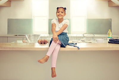Buy stock photo Portrait of a cute young mixed race girl sitting on the kitchen counter with an apron and smiling. Little hispanic girl smiling and sitting alone while baking home. She enjoys cooking in the kitchen