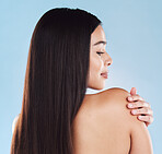 One beautiful young hispanic woman with healthy skin and sleek long hair looking over and touching shoulder while posing against a blue studio background. Mixed race model with flawless complexion and natural beauty
