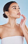 Beautiful young mixed race woman touching her mouth and posing isolated in studio against a blue background. Attractive female looking confident and happy with her daily skincare regime