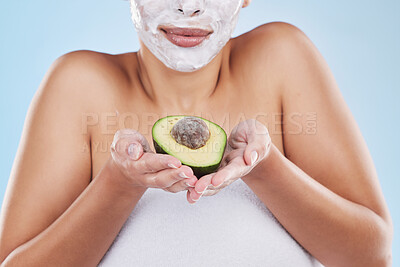 Beautiful young mixed race woman wearing a face mask peel and posing with an avocado isolated in studio against a blue background. A skincare regime keeps her fresh. Packed with vitamins and nutrients