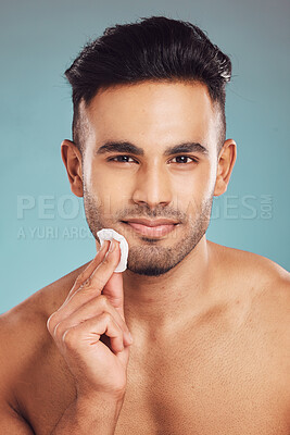 Buy stock photo Portrait of one young indian man wiping a round cotton swab on his face while grooming against a blue studio background. Handsome mixed race guy cleaning and exfoliating his face for a healthy complexion and clear skin