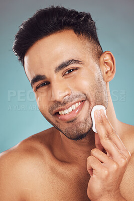 Buy stock photo Portrait of one smiling young indian man wiping a round cotton swab on his face while grooming against a blue studio background. Handsome mixed race guy cleaning and exfoliating his face for a healthy complexion and clear skin
