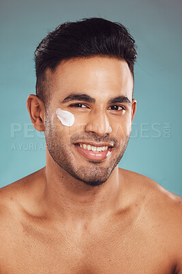 Buy stock photo Portrait of one smiling young indian man applying moisturiser lotion to his face while grooming against a blue studio background. Handsome guy using sunscreen with spf for uv protection. Rubbing facial cream on cheek for healthy complexion and clear skin