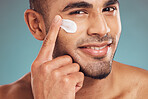 Portrait of one smiling young indian man applying moisturiser lotion to his face while grooming against a blue studio background. Handsome guy using sunscreen with spf for uv protection. Rubbing facial cream on cheek for healthy complexion and clear skin