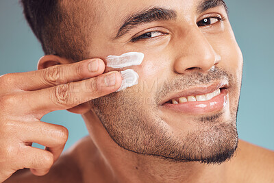 Portrait of one smiling young indian man applying moisturiser lotion to his face while grooming against a blue studio background. Handsome guy using sunscreen with spf for uv protection. Rubbing facial cream on cheek for healthy complexion and clear skin