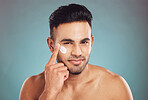 Portrait of one young indian man applying moisturiser lotion to his face while grooming against a blue studio background. Handsome guy using sunscreen with spf for uv protection. Rubbing facial cream on cheek for healthy complexion and clear skin