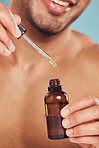 Closeup of one mixed race man using a dropper to apply serum oil from a bottle to his skin and face against a blue studio background. Guy holding a moisturising aftershave product for healthy, smooth and soft skin