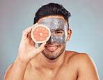 Portrait of one handsome young indian man applying a detoxifying clay or charcoal facial mask while holding grapefruit against a blue studio background. Guy using moisturising products with natural ingredients on his face for healthy, smooth soft skin