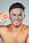 Portrait of one handsome young indian man applying a detoxifying clay or charcoal facial mask while holding grapefruit against a blue studio background. Guy using moisturising products with natural ingredients on his face for healthy, smooth soft skin