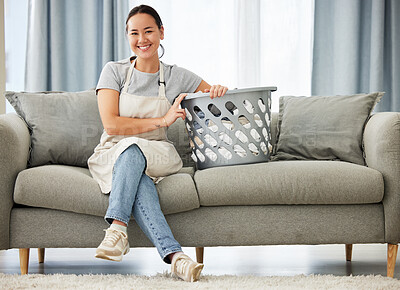 Portrait of a beautiful young mixed race woman holding a laundry basket while cleaning her apartment. One happy Asian woman smiling while folding laundry at home