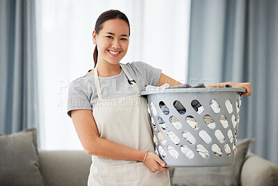 Portrait of a beautiful young mixed race woman holding a laundry basket while cleaning her apartment. One happy Asian woman smiling while folding laundry at home