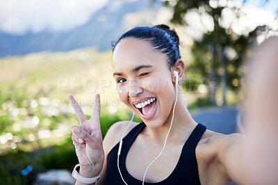 Portrait of one fit young mixed race woman listening to music with earphones and gesturing peace sign for selfies while taking a break from exercise outdoors. Happy female on video call and taking photos for social media during a rest from running workout