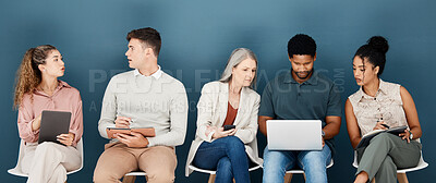 Group of diverse serious businesspeople sitting on chairs in a row in an office together. Five business professionals sitting and talking using technology in a waiting room together in an office