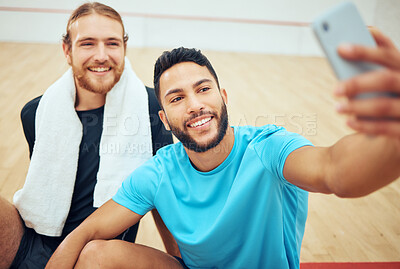 Two young athletic squash players using cellphone for selfie after playing court game. Smiling fit active Caucasian and mixed race athlete sitting together and feeling happy while posing for picture