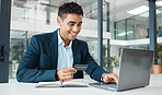 Young happy businessman holding and using a credit card and typing on a laptop alone at work. One content hispanic male businessperson smiling while making an online purchase with his debit card and laptop