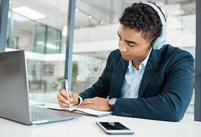 Buy stock photo Young serious mixed race businessman wearing headphones listening to music while writing ideas in a notebook alone at work. One hispanic male businessperson planning in a diary in an office