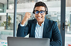 Young happy mixed race male call center agent answering calls while wearing a headset and glasses working on a laptop alone at work. One hispanic businessperson smiling while cheering at a desk in an office