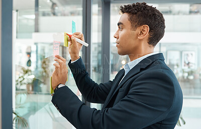 Buy stock photo Serious mixed race businessman writing on a note planning ideas on a glass window alone at work. One focused hispanic businessperson brainstorming in an office