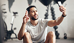 One fit young hispanic man listening to music with earphones from a cellphone and gesturing a peace sign for selfies while taking a break from exercise in a gym. Happy guy making video call and taking photos for social media during a rest from workout