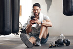 One fit young hispanic man listening to music with earphones from a cellphone while taking a break from exercise in a gym. Happy guy texting, making video call and using fitness apps online while browsing social media and watching workout tutorials