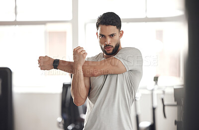 Portrait of one fit young hispanic man stretching arms and shoulders for warmup to prevent injury while exercising in a gym. Serious muscular guy focused on staying motivated and determined while mentally and physically preparing for training workout