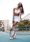Cheerful young girl standing on the tennis court. African american tennis player enjoying her practice outside. Young athlete holding her tennis racket next to the net at her club
