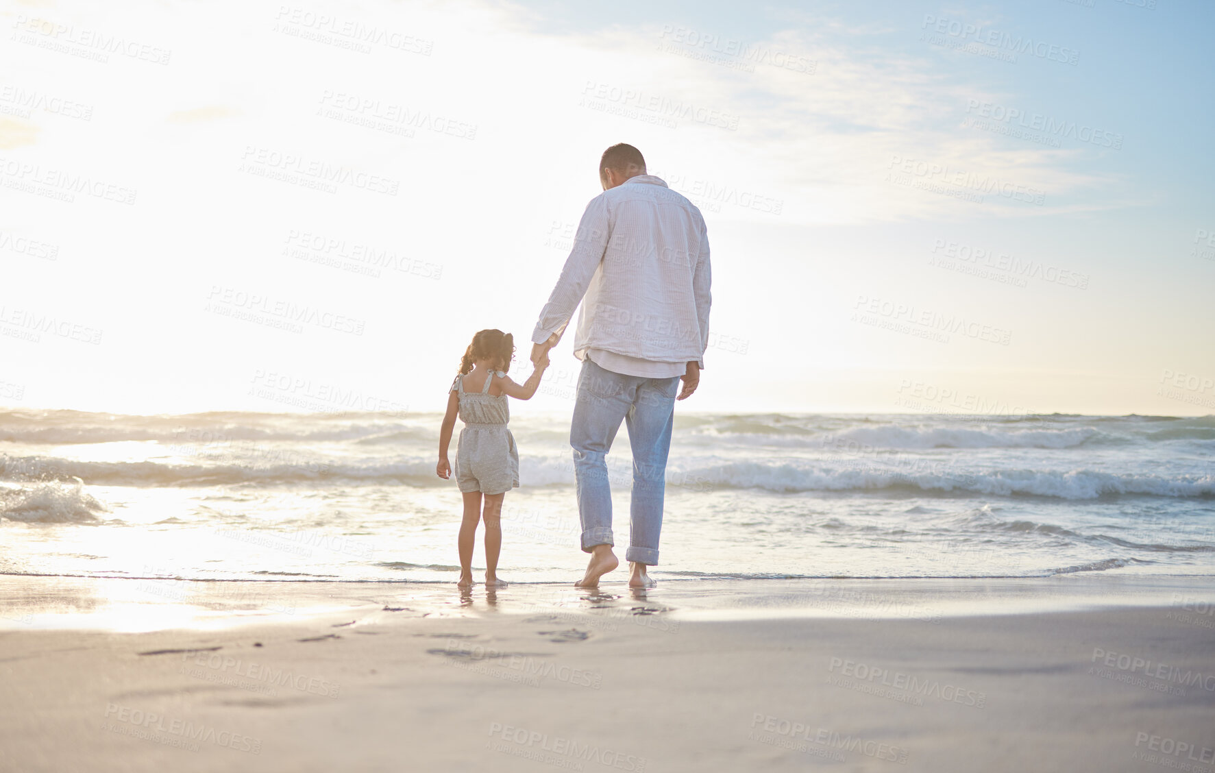 Buy stock photo Rearview cute mixed race girl standing hand in hand with her father in the sea at the beach. A young man and his daughter holding hands while standing in the water and looking at the view at sunset