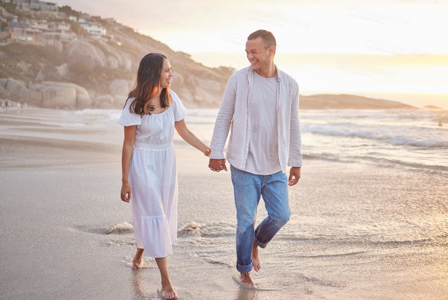 Buy stock photo Affectionate mixed race couple holding hands and walking along the beach. Husband and wife hand in hand in the sand by the sea. Enjoying a romantic walk by the seashore at sunset. Love is in the air