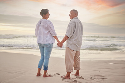 Buy stock photo Rearview affectionate mature mixed race couple standing hand in hand on the beach. Senior husband and wife holding hands by the sea. They love spending time together on the coast in summer at sunset
