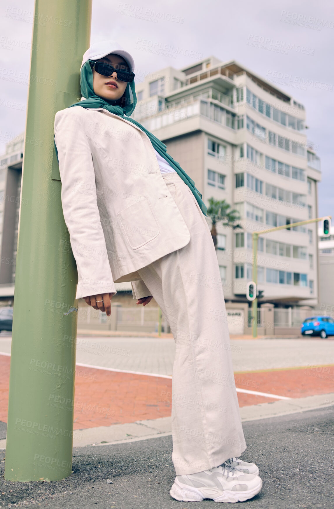 Buy stock photo Portrait, city fashion or religion with a muslim woman leaning against a pole in a cap and scarf for contemporary style. Islam, faith or hijab with a trendy young arab person outside in an urban town
