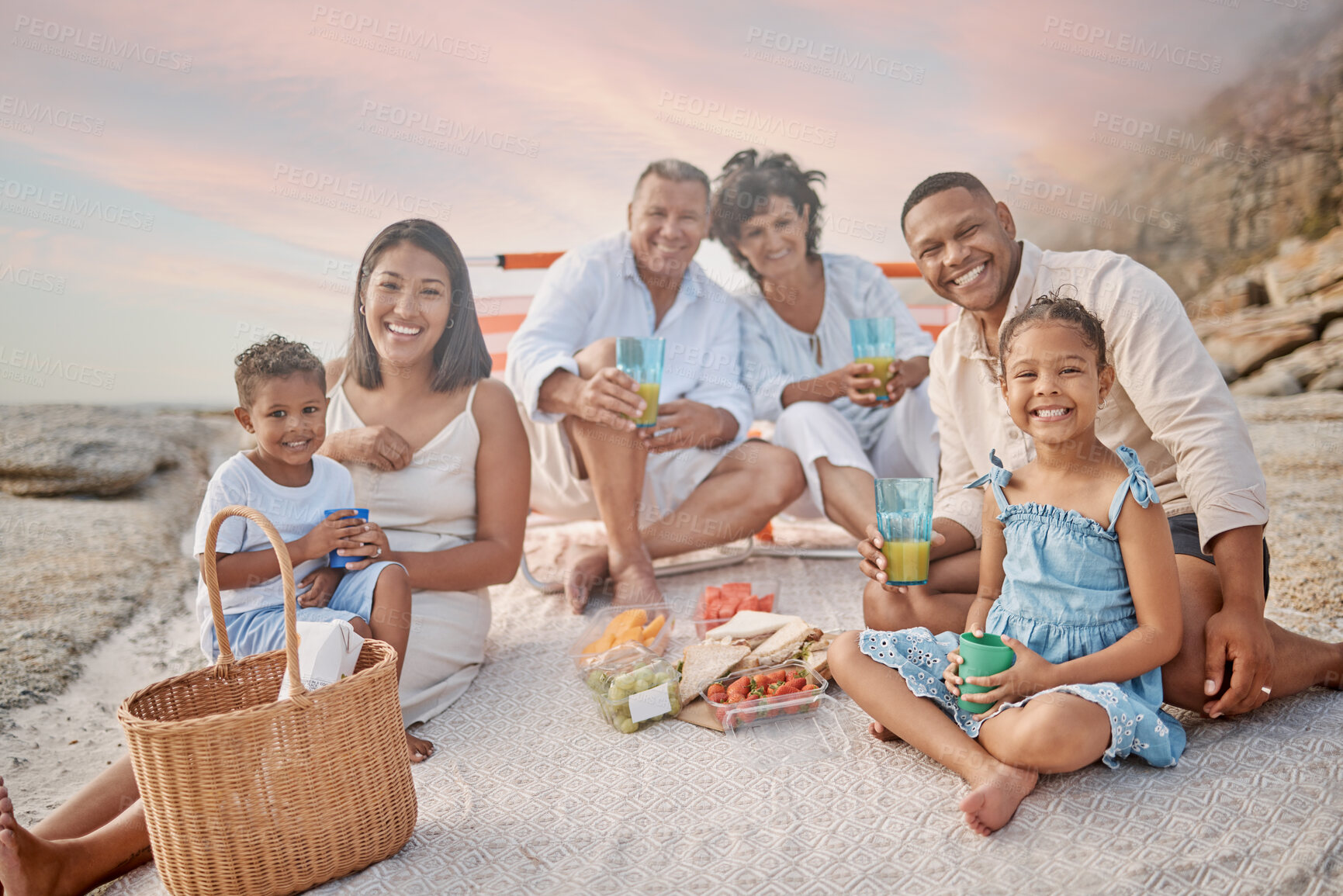 Buy stock photo Closeup of a mixed race family having a picnic on the beach and smiling  while having some food with snacks. Happy family bonding on a day out at the beach