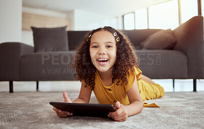 Portrait of adorable little mixed race child using digital tablet in home living room. One small cute hispanic girl lying alone on lounge floor, playing game on technology. Happy kid with curly hair