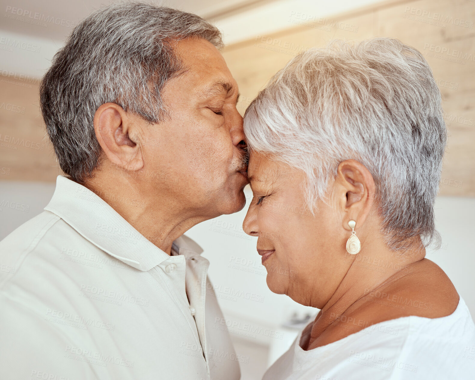 Buy stock photo Closeup of mixed race senior man kissing wife’s forehead in the morning at home. Affectionate elderly husband and wife standing and bonding in kitchen. Two happy retired man and woman feeling in love