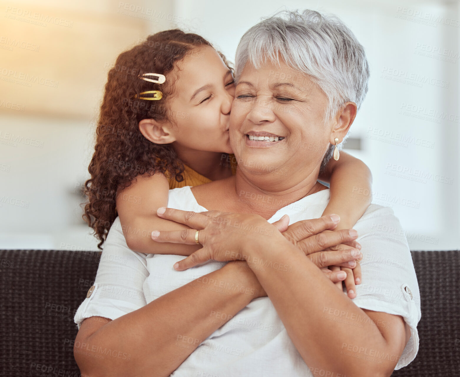 Buy stock photo Mixed race granddaughter kissing grandmother’s cheek in living room at home. Senior woman enjoying weekend with adorable little girl in lounge. Playful hispanic child bonding and hugging grandparent