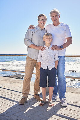 Buy stock photo Male family members posing together at the beach on a sunny day. Grandfather, father and grandson standing together on seaside promenade. Multi-generation family of men and little boy spending time together