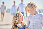 Happy little girl and boy holding hands and talking while walking together on seaside promenade on a sunny day while their parents follow in the background. Cute sibling sister and big brother getting along