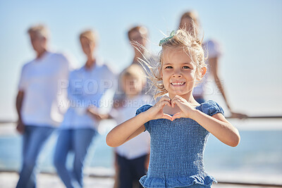 Buy stock photo Portrait of an adorable little girl showing a heart shape with her hands with her family standing in the background. Cute kid having fun at the beach on a sunny day