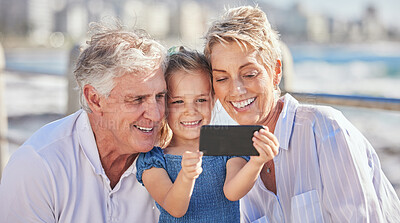 Buy stock photo Cute little caucasian girl smiling while holding mobile phone and taking a selfie or video call with her grandparents. Happy senior grandmother and grandfather posing for a photo with their granddaughter outdoors on a sunny day