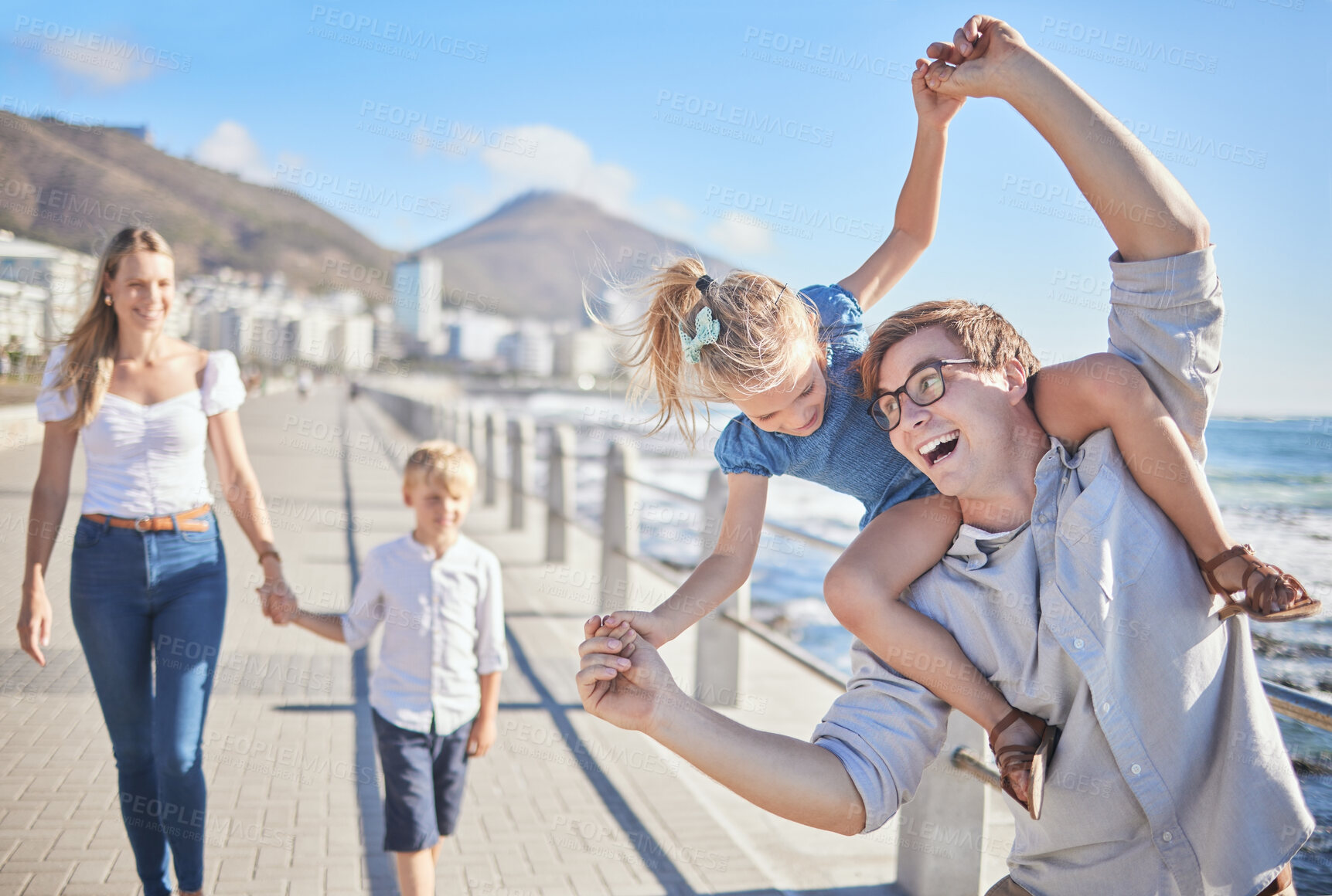 Buy stock photo Cheerful caucasian father carrying his daughter on his shoulders being playful and having fun while his wife and son follows in the background on a seaside promenade