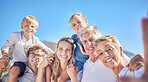 Portrait of happy caucasian multi-generation family standing together and taking a selfie outdoors against a blue sky. Two little children sitting on their parents shoulders while enjoying time at the beach with their parents and grandparents against a bl
