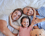 Adorable little girl pulling her parents close while lying in between her mother and father, from above. Faces of loving parents bonding with their daughter lying on a bed and spending free time together on the weekend