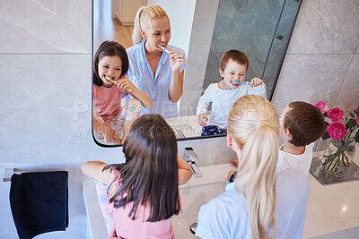 Above view of caucasian family using toothbrushes and looking in mirror. Young caucasian mother in pyjamas standing with her children while they brush their teeth in the bathroom at home. Little sibling brother and sister doing morning routine with mom te