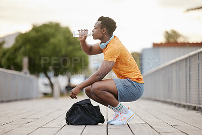 An African American man drinking water on a bridge. A man taking a break from exercising drinking from a bottle. A sporty guy focusing on health.